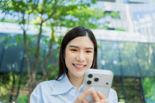 Close portrait of a smiling asian woman looking at her smartphone screen outdoors, framed by green leaves and a modern glass building.