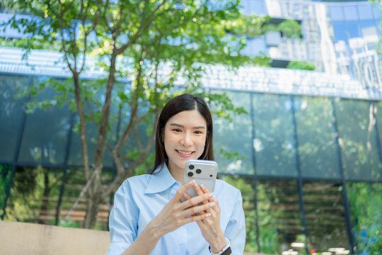 Asian businesswoman types a message on her smartphone with both hands while standing outdoors under green trees beside a glass office building.