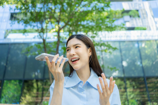 Young Asian woman speaks into her smartphone to record a voice message while standing outside among green trees, gesturing with her free hand as she talks.