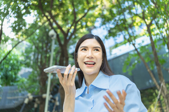 Low angle shot of an asian businesswoman laughing while sending a voice message on her smartphone under a canopy of green leaves.