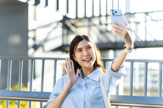Backlit asian woman takes a selfie and waves at her smartphone camera on an elevated city skywalk in warm evening sunlight.