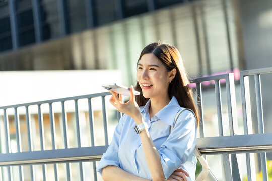 Asian businesswoman leans against a railing and speaks through a smartphone speaker, bathed in soft sunlight in the business district.