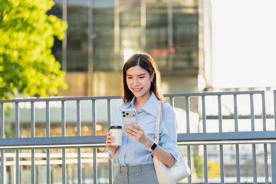 Asian businesswoman holds a takeaway coffee cup and reads her smartphone while walking along a city street on her morning commute.