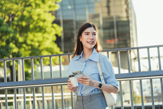 Asian businesswoman holds a coffee cup and a smartphone and looks away with a smile outdoors, with open space for text on the left.