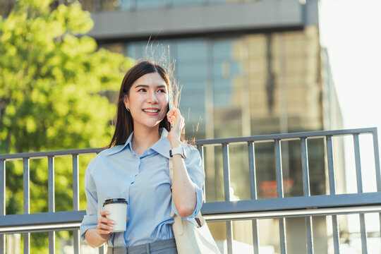 Asian businesswoman talks on her mobile phone and holds a coffee cup outdoors, her hair moving in the wind on a morning city commute.