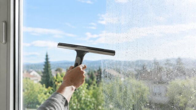 Hand using a squeegee to clean a window, revealing a view of trees and hills under a clear blue sky in a residential area