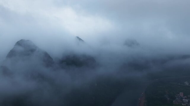 Misty karst mountain peaks in Guilin emerge through dense fog, with a river and small village visible below under overcast daylight.