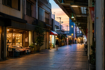 Fototapeta premium Dusk Falls on the Shopping Arcade Warm shop lights glow along a quiet covered arcade as twilight settles over the rooftops, with a few passersby heading home.
