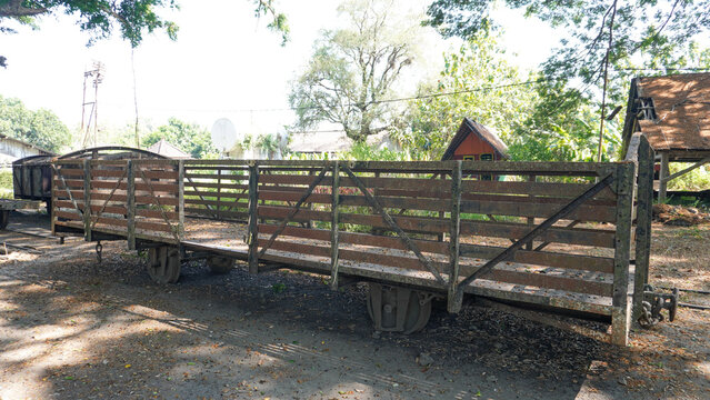 An old rusty teak wood train wagon under a large shady tree near a traditional wooden shed in Cepu, Central Java, Indonesia, showcasing historic industrial railway forestry heritage in rural nature.