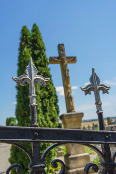 Ornate wrought iron fence spearheads in foreground with crucifix and green cypress tree in background under blue sky, religious monument detail