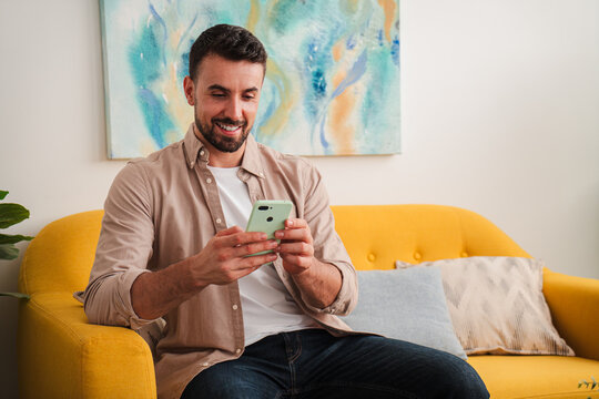 A friendly man sits on a bright yellow sofa, focused on a smartphone, checking messages, browsing apps, and enjoying a lazy afternoon. The calm living room mood shows everyday connectivity in action..
