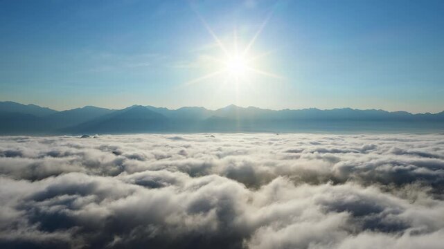 Wide sea of clouds covers the valley below distant mountain ridges, with a bright sun shining in the clear blue sky.