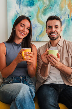 Two people sit close together holding mugs and smiling toward camera, conveying friendly morning ritual, relaxed mood, warm colors, and casual comfort in a candid portrait. Warm light highlights Joy!