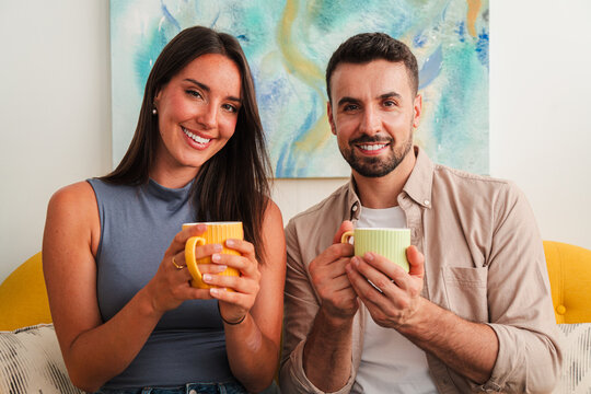 A joyful pair sits close on a sofa, holding colorful cups while sharing a cheerful moment. The scene emphasizes warmth, companionship, and relaxed comfort during a quiet morning together.