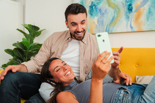 A smiling couple lounges on a bright couch and uses a sleek phone to snap a spontaneous selfie, conveying warmth, affection, and everyday companionship in a cozy living space.