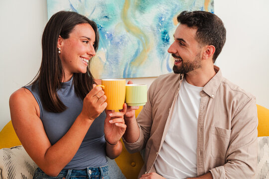 A cheerful couple raises bright mugs in a friendly toast, eyes smiling as they enjoy a cozy moment together on a soft sofa during a quiet morning at home