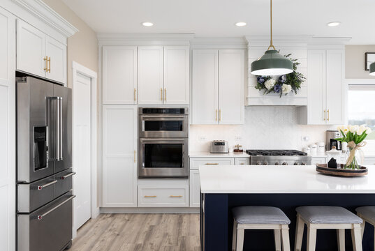 A white kitchen with green lights hanging over a blue island, a white marble countertop, stainless steel appliances, and a chevron tile backsplash. No brands or labels.