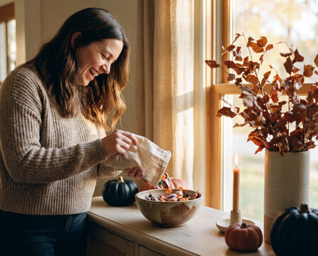 Smiling woman pouring halloween candy into ceramic bowl on cozy autumn kitchen counter