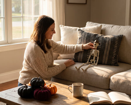 Smiling woman positioning small toy skeleton wearing hat on sofa pillow at halloween home