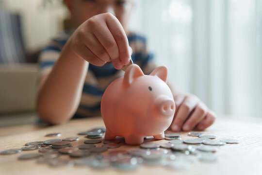Asian boy inserting a coin into a piggy bank surrounded by coins at home, representing saving money, financial education, budgeting, responsibility, future planning and childhood learning.