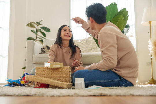 Happy Asian couple unpacking and organizing belongings in their new home while sitting together on the floor, representing moving, relocation and homeownership.