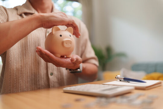 Man holding a piggy bank above a calculator and coins at home, representing saving, financial planning, budgeting, emergency funds, security, investment and future financial stability.