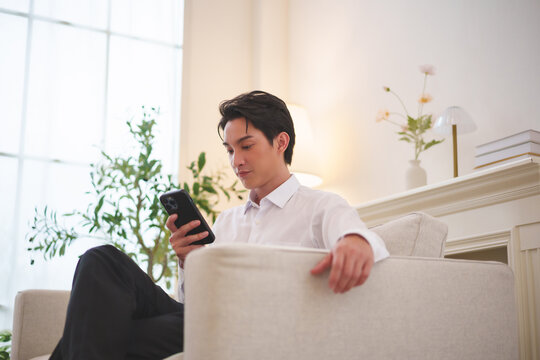 Asian man relaxing on a sofa while using a smartphone at home, representing mobile lifestyle, online browsing, communication, entertainment, leisure time and everyday digital convenience.