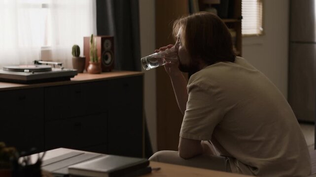 Caucasian young adult man sitting indoors, drinking fresh water from glass and smiling thoughtfully. Clip suitable for healthcare campaigns, hydration awareness or daily wellness projects