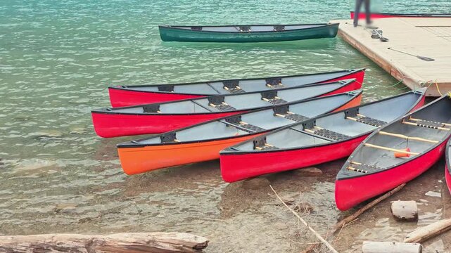 Multiple vibrant canoes are resting by the clear turquoise waters, reflecting in Lake Moraine, inviting visitors to enjoy summer outdoor recreation and explore Banff National Park in Alberta, Canada