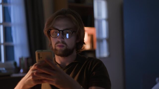 Young adult Caucasian man with glasses using smartphone in dark room at night, browsing online content. Ideal for projects on digital lifestyle, mobile applications, and insomnia awareness