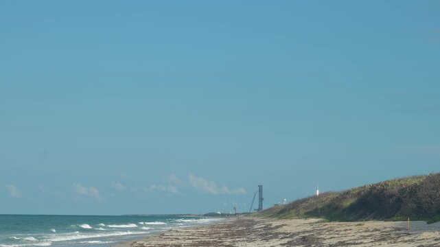 A rocket lifts into a clear blue sky above a sandy beach and ocean shoreline. The coastal spaceport, launch tower, waves, dunes, and bright daylight create a dramatic aerospace travel scene.