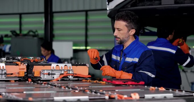 Adult male technician handles battery component and checks digital tablet beside EV battery pack in automotive workshop, combining technical inspection, diagnostics, preventive maintenance service.