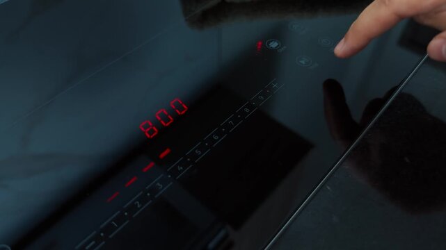 Person's finger interacting with a sleek black glass induction cooktop, setting the power level with illuminated digital controls and displaying the chosen heat intensity for cooking