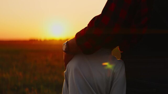 Warm sunset moment of family bonding as an adult gently rests a comforting hand on a child shoulder while watching the glowing horizon over a peaceful field