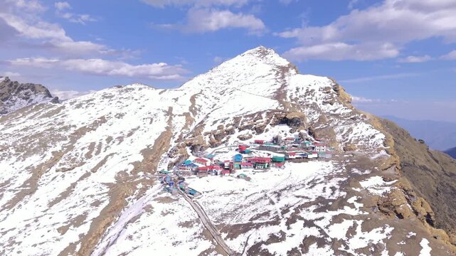 Aerial drone view of Tungnath Mahadev Temple in Chopta, Uttarakhand, India, surrounded by snow-covered Himalayan mountains during winter. The footage shows the ancient Shiva temple, nearby local marke