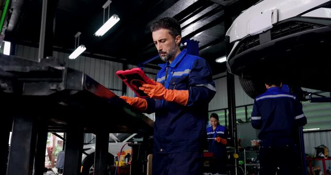 Adult male technician checks digital tablet beneath raised electric vehicle in modern automotive workshop, supporting vehicle diagnostics, undercarriage inspection and preventive maintenance.
