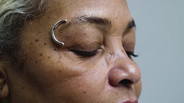 Blinking senior woman lowering right eyelid in studio, showing brow ring and false lashes