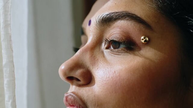 Blinking Indian woman closing lids at window as sunlight hitting eye showing purple bindi gold stud