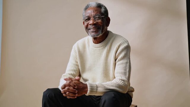 Pulling back cam, older man smiling gesturing clasping posing portrait in set wearing sweater specs