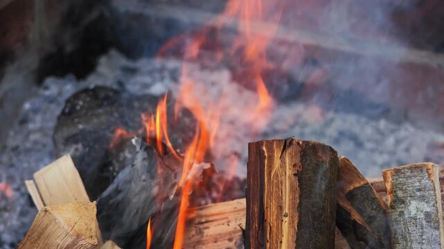 Slow-motion video of bright orange flames rising and twisting above burning firewood in an outdoor brick fire pit. Smoke drifts through the frame while the flames move dynamically against the dark bac