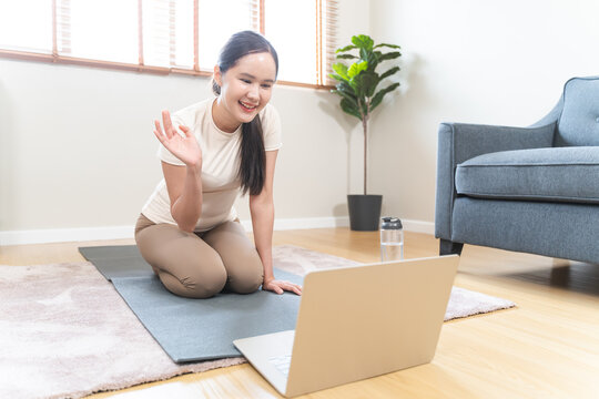 Active asian young woman practicing yoga on mat looking at laptop computer screen watching online fitness class at home living room for wellness lifestyle and self care