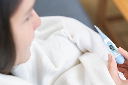 Close-up of sick Asian woman hand holding a digital thermometer showing high body temperature of 39.7 degrees celsius, female wrapping in white blanket having a severe fever at home