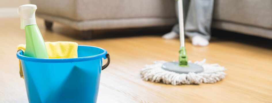 Close up of a blue bucket with cleaning spray bottle and microfiber cloth on the wooden laminate floor with a person mopping the living room in the background during daily domestic chores routine.