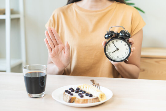 Asian young woman raising hand in stop gesture refusing healthy breakfast food while holding alarm clock, intermittent fasting weight loss regimen and time restricted eating nutrition concept.