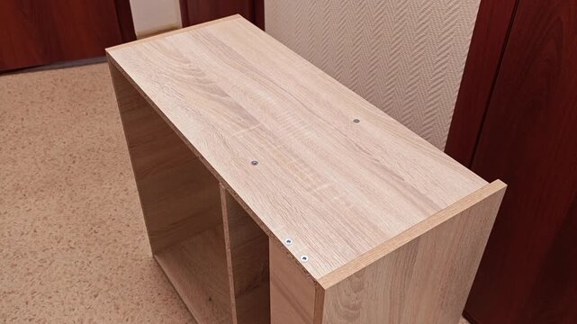 Wooden shelf unit fills frame in empty room. Shelf cabinet stands beside patterned wall and closed door, with camera moving closer to highlight unfinished top panel, screw heads, and light wood grain.