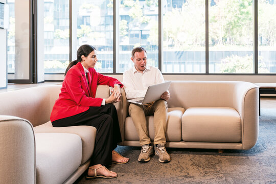 Two colleagues on a modern office sofa review work on a laptop near large windows, collaborating on a project in a bright, contemporary lounge.