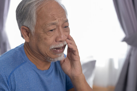 Elderly asian man suffering from severe toothache pain holding hand to jaw in deep distress standing indoor near bright daylight window