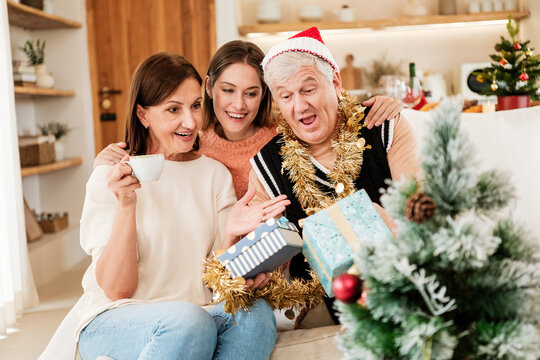Three adults smile and share wrapped Christmas gifts beside a festive tree at home, enjoying a cheerful holiday moment filled with surprise, warmth and togetherness.