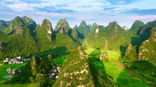 Lush green karst mountains rise above agricultural fields and a small village in Guilin, illuminated by bright sunlight under a blue sky.