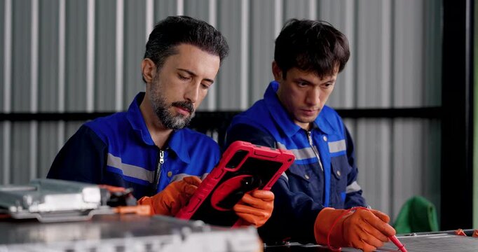Two male technicians test EV battery system using digital tablet, meter and probe in automotive workshop, combining battery diagnostics, electrical inspection, teamwork and technical maintenance.